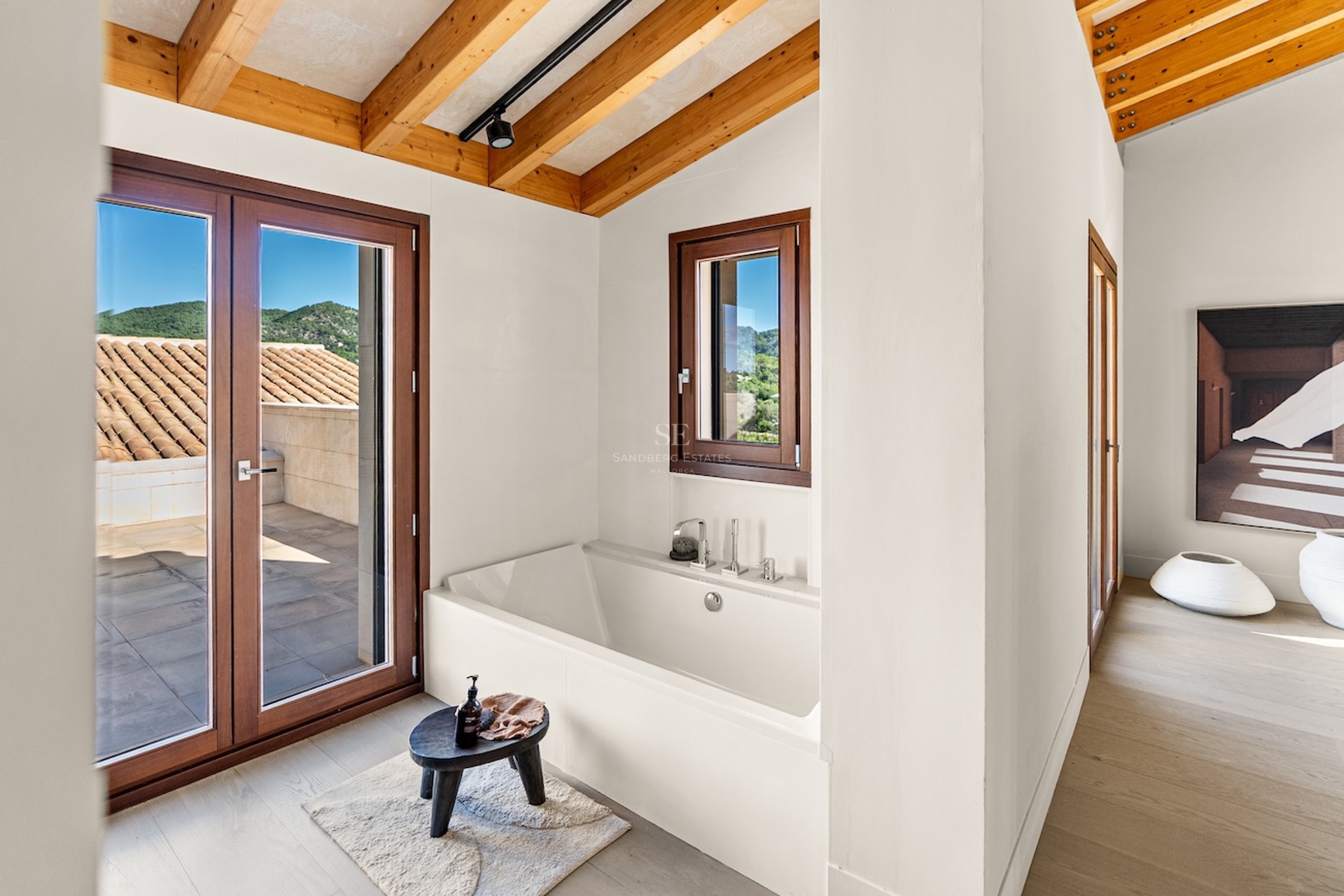 Baignoire blanche sous un plafond en poutres de bois avec accès terrasse et vue sur les montagnes.