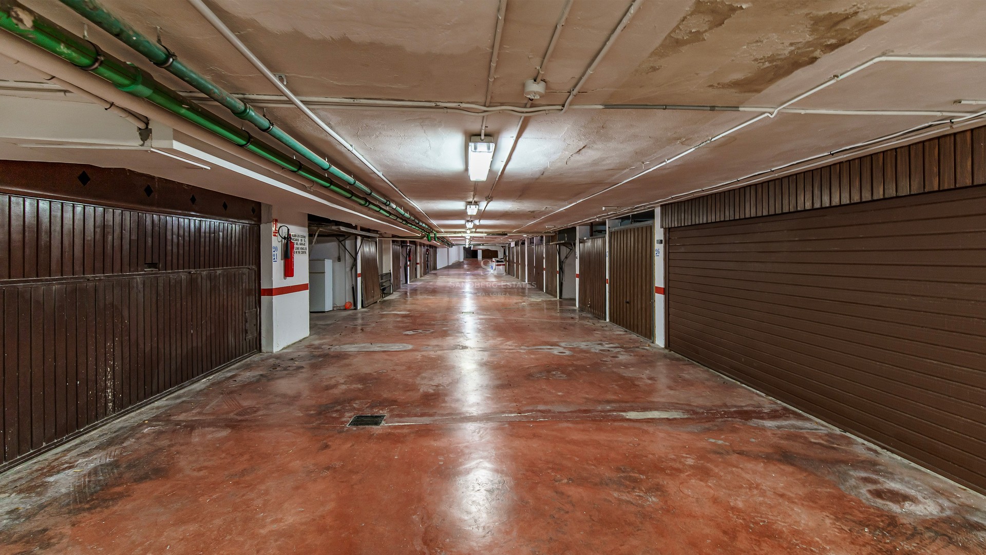 Underground garage hallway with red concrete floor and individual brown garage doors on both sides.