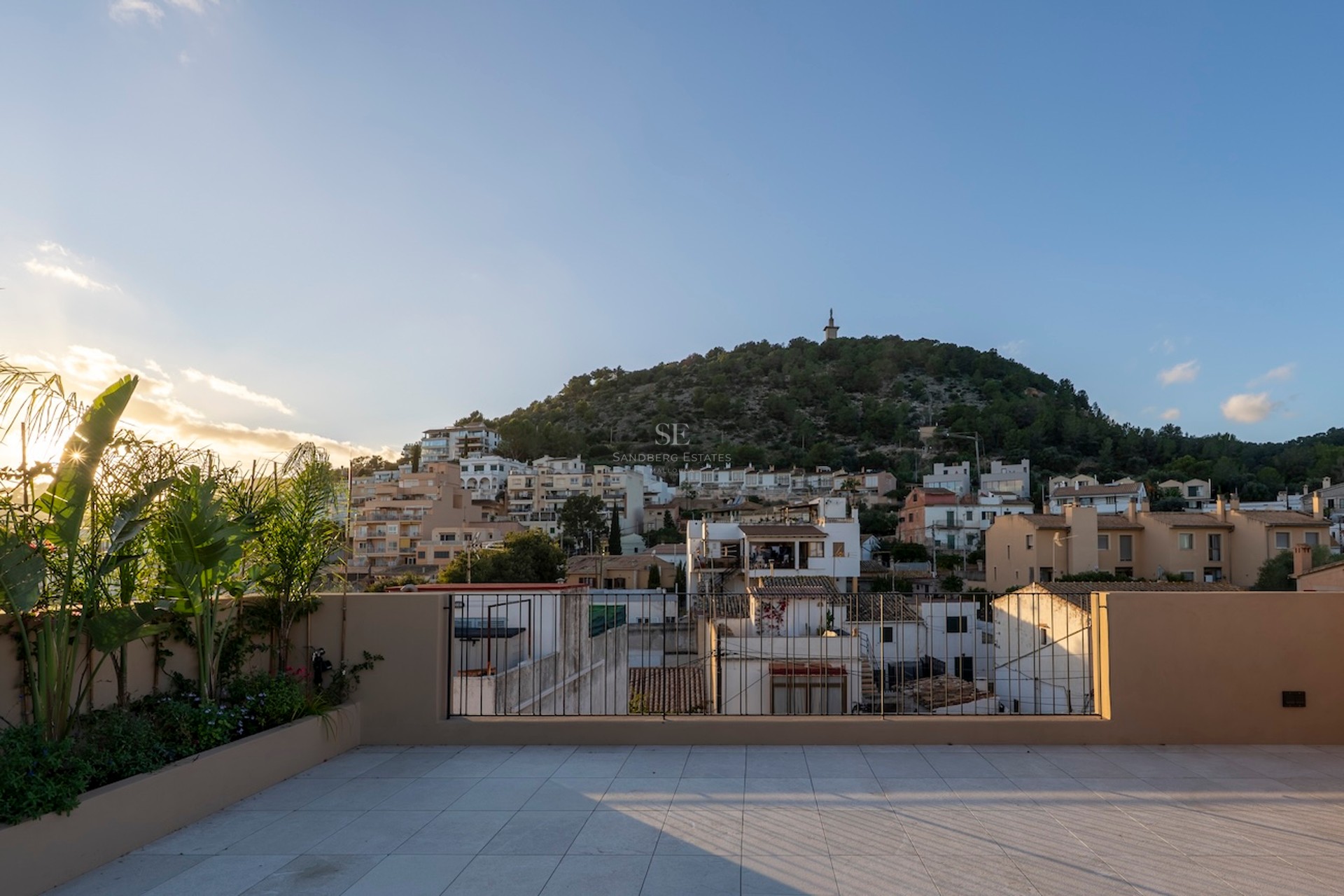 Grande terrasse carrelée avec garde-corps métallique offrant une vue sur un village méditerranéen et une colline au coucher du soleil.