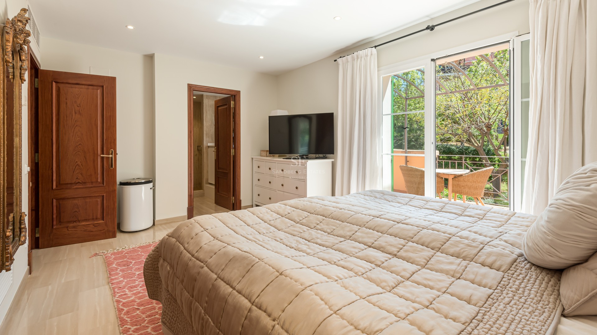 Bright bedroom featuring quilted bedding, wooden doors, and sliding glass doors to a terrace with wicker furniture.