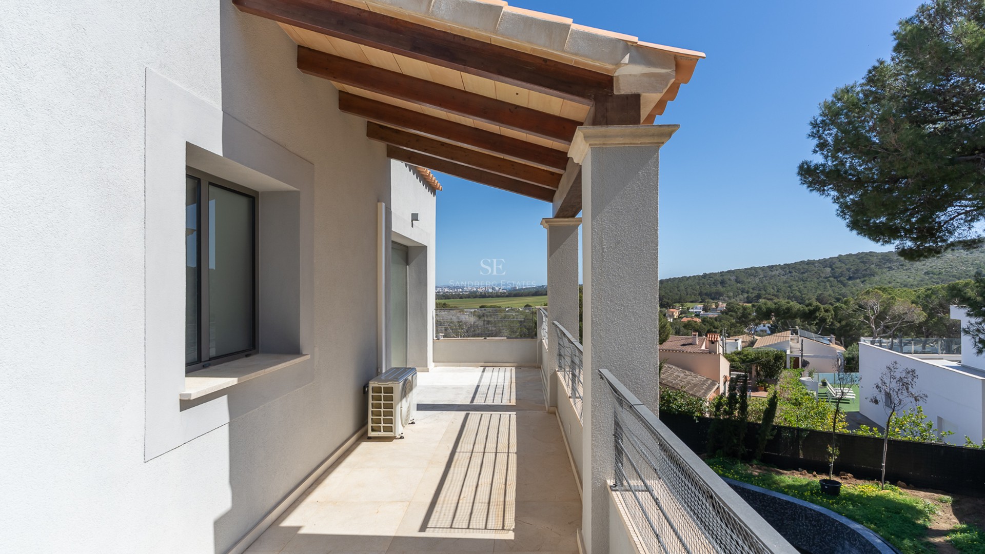 Lång övertäckt balkong med träbjälkar, vita väggar och metallräcke med utsikt över en grönskande dal.