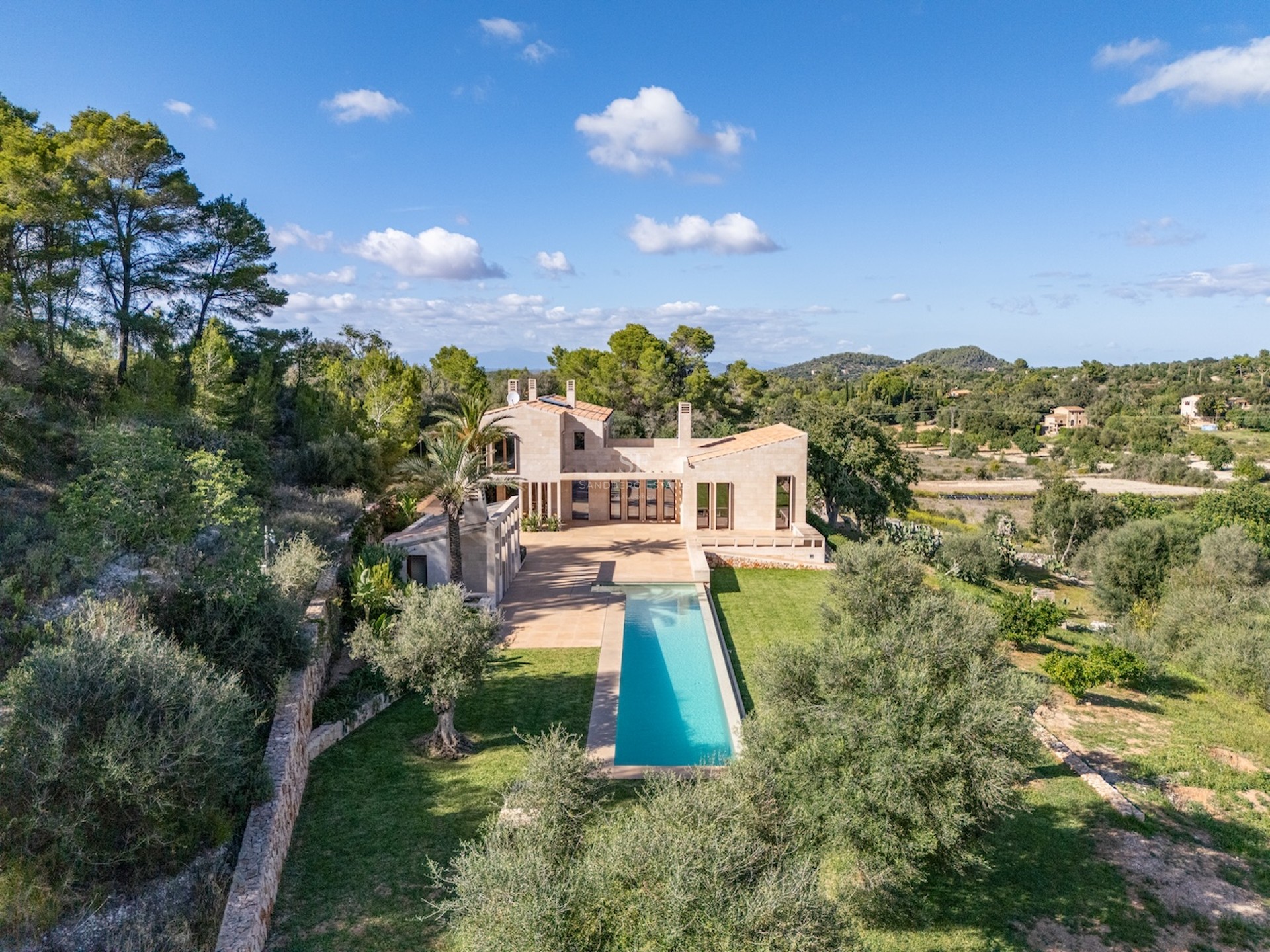 Vue d'une villa moderne en pierre avec une longue piscine turquoise entourée d'une végétation méditerranéenne luxuriante.