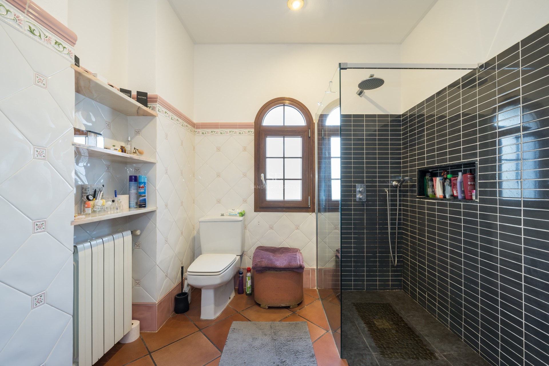 Bathroom featuring terracotta floors, decorative wall tiles, an arched wooden window, and a modern glass walk-in shower.