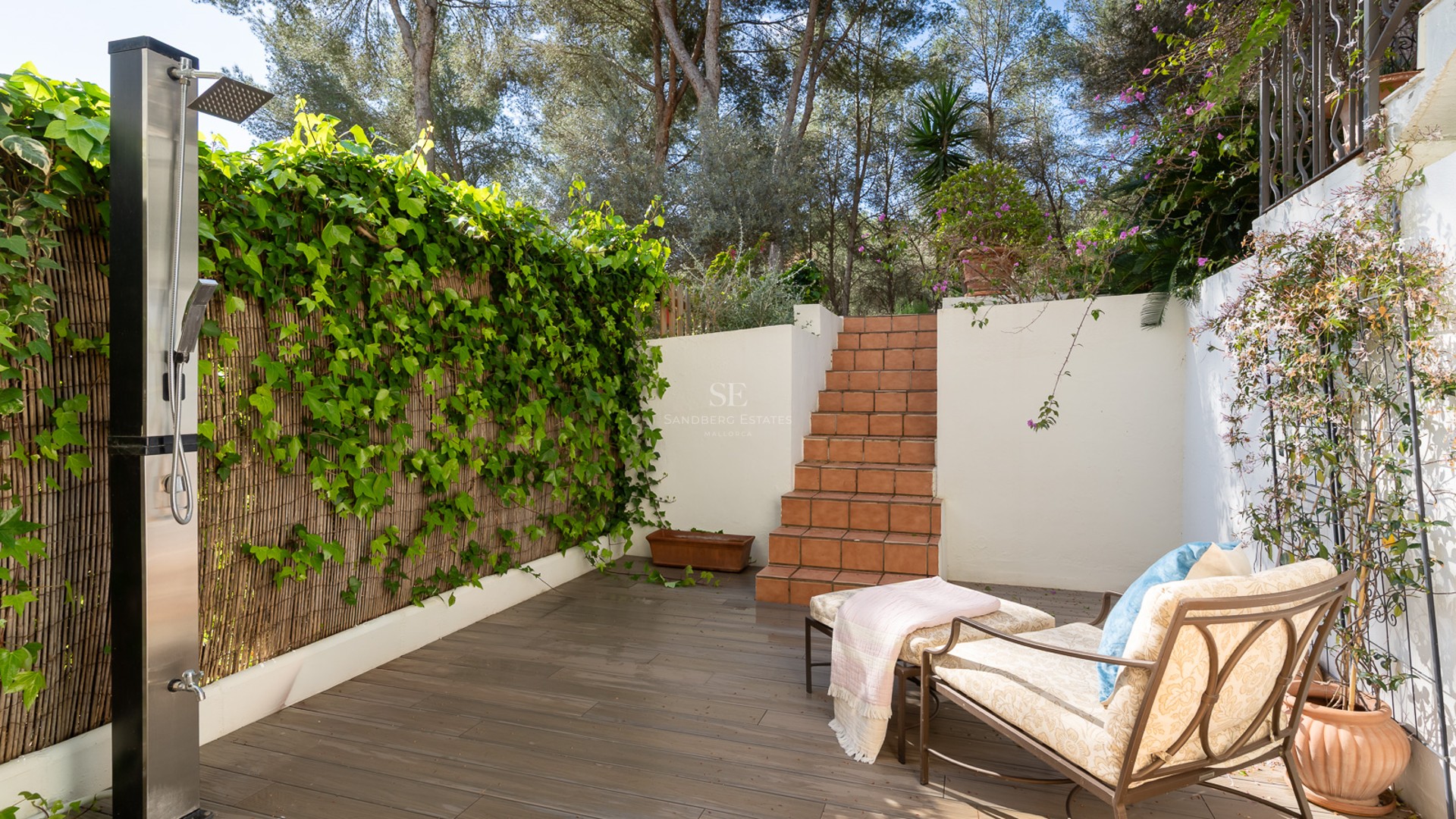 An outdoor terrace featuring a modern stainless steel shower, terracotta steps, a lounge chair, and a green ivy wall.