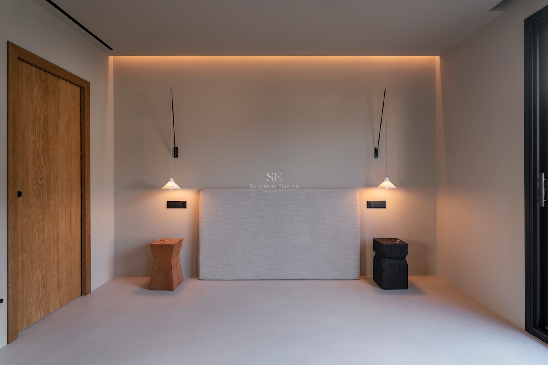 Tête de lit rembourrée entre des suspensions et des tabourets en bois sous un éclairage chaleureux et indirect.