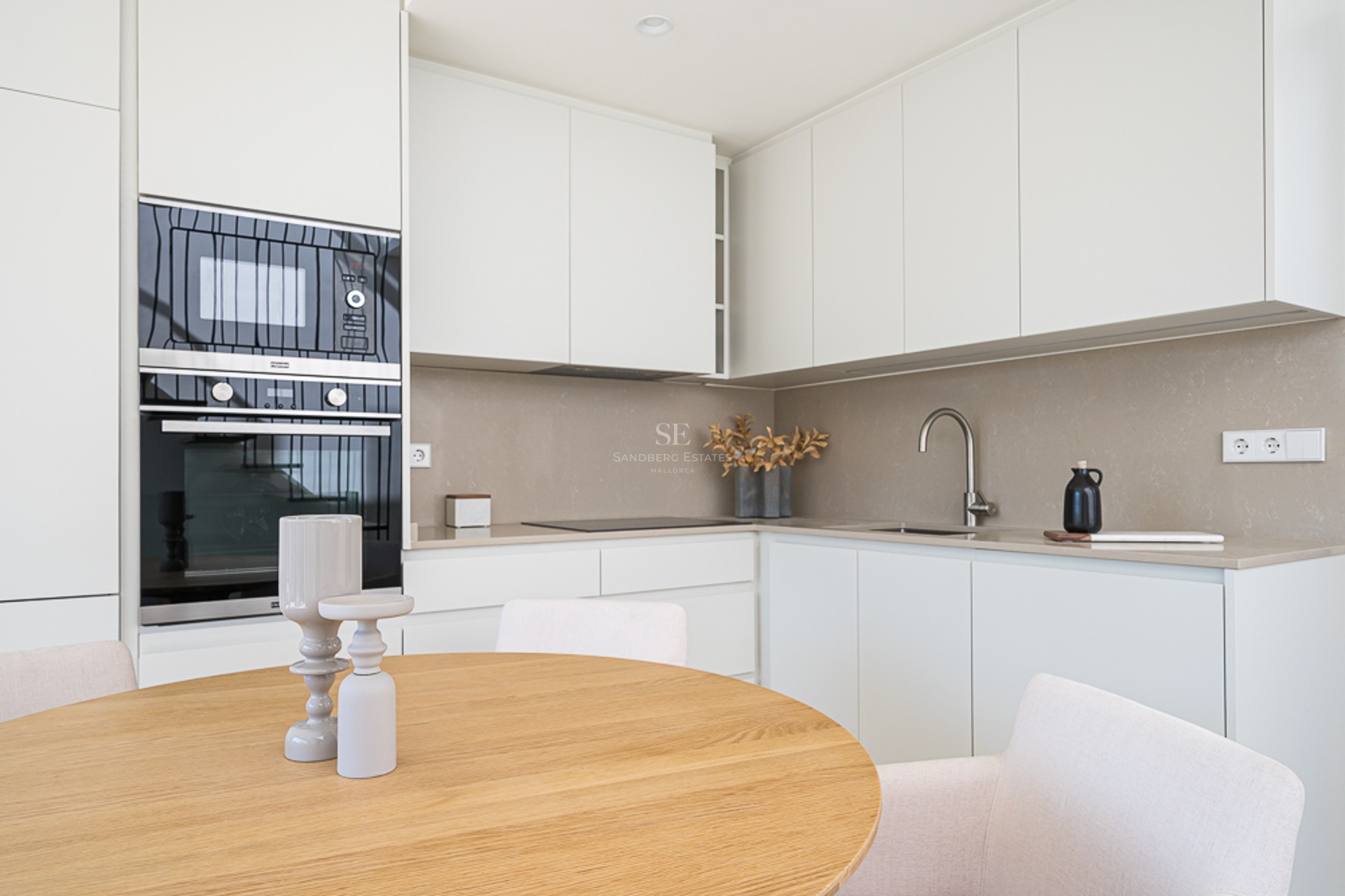 Contemporary white kitchen featuring matte cabinets, stone countertops, and a wooden dining table in the foreground.