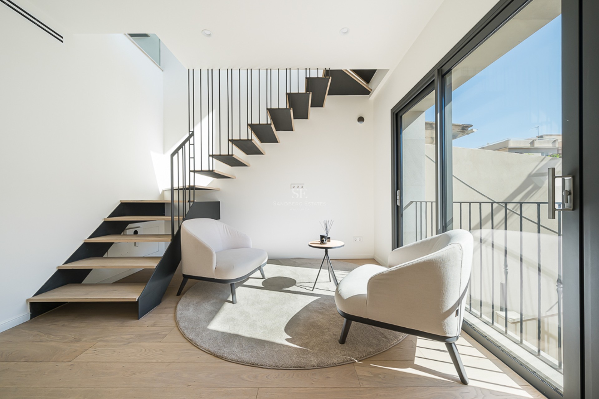Modern living space featuring a floating wooden staircase, two cream armchairs, and floor-to-ceiling glass doors.