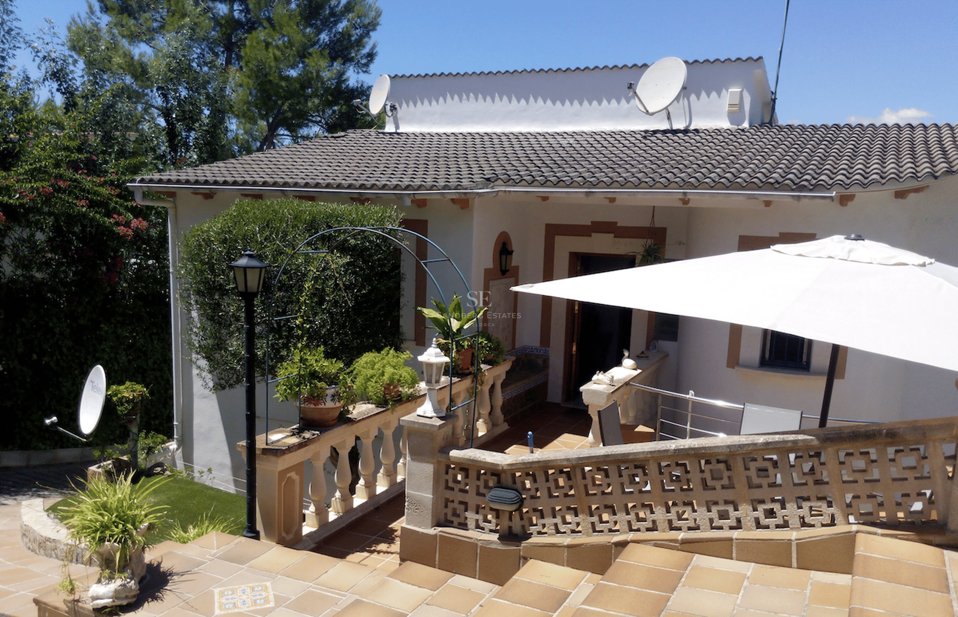 Terraza de terracota con balaustrada de piedra, sombrilla blanca y vegetación bajo un cielo despejado.