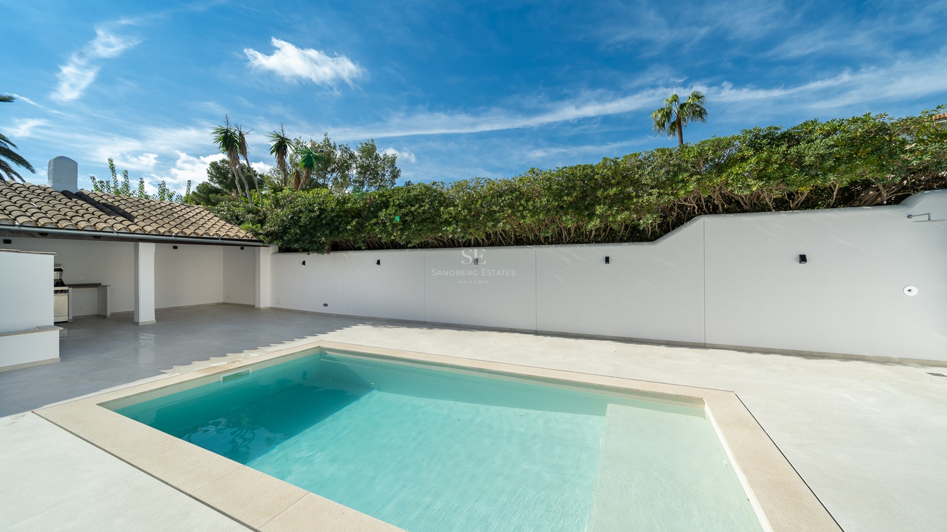 Piscine turquoise sur terrasse blanche avec mur d'intimité, haies vertes et cuisine d'été sous ciel bleu.