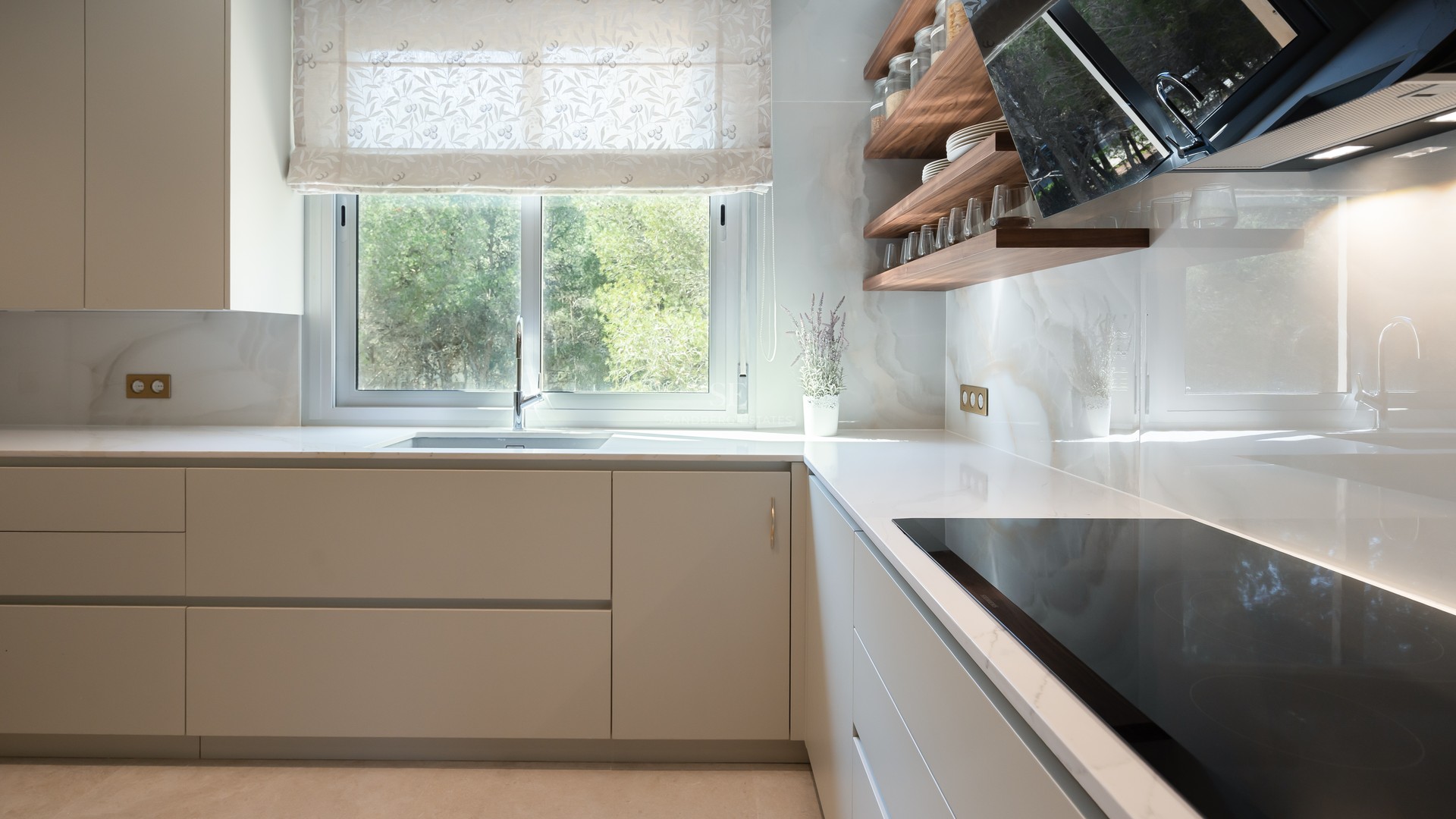 Minimalist off-white kitchen with marble-effect backsplash, wooden shelves, and a window overlooking lush greenery.