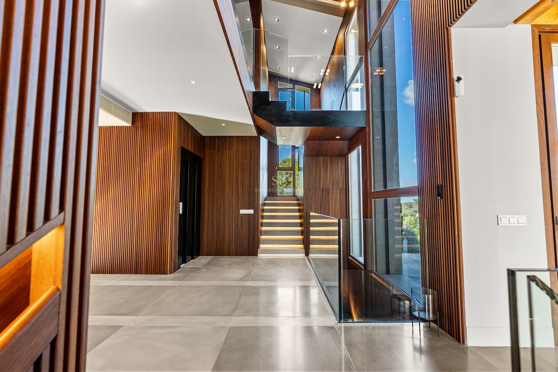 Hall d'entrée moderne avec murs en bois à lattes, escalier en verre avec éclairage LED et grandes baies vitrées.
