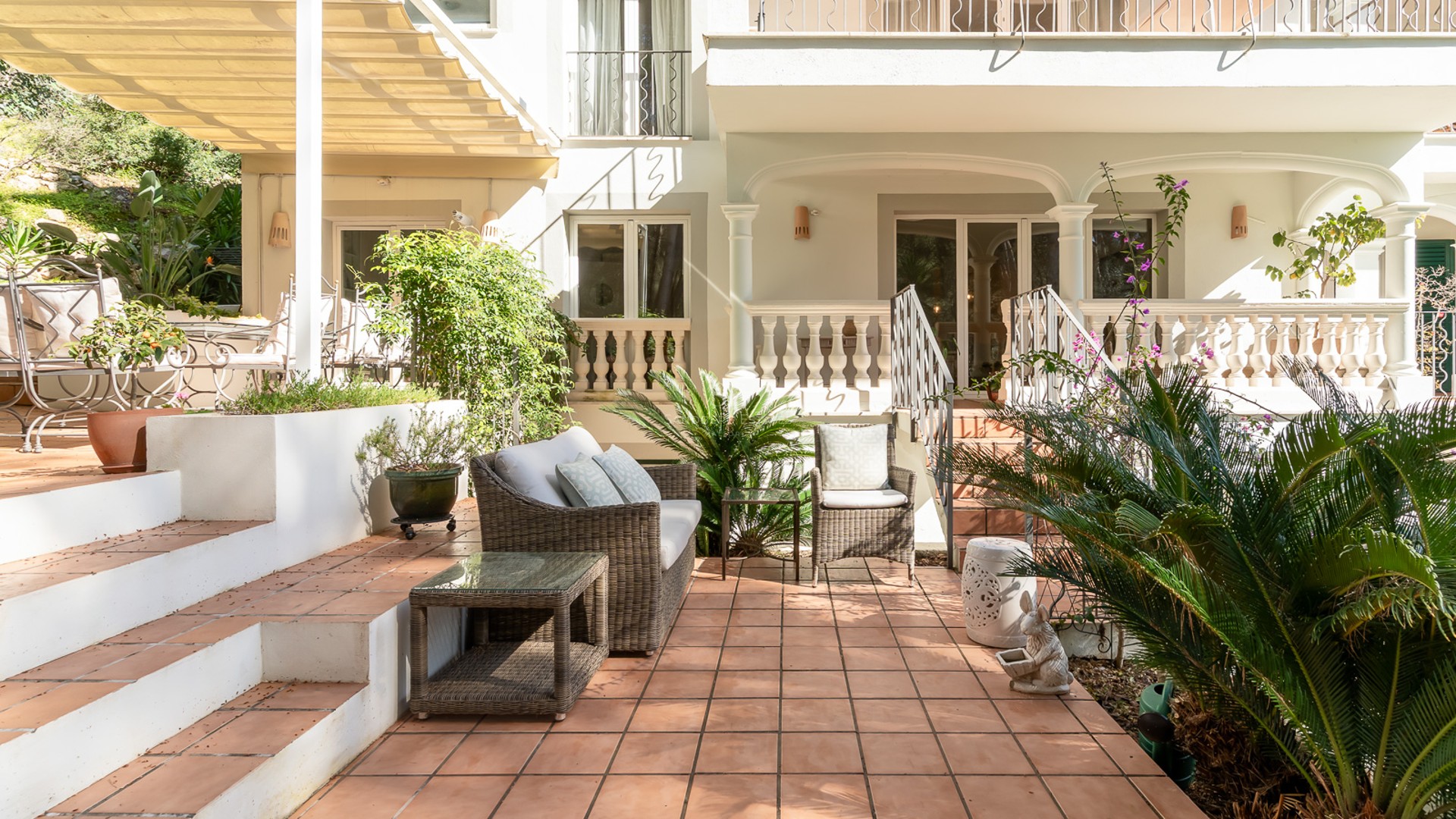 Sun-drenched terracotta tile terrace with wicker lounge furniture and lush greenery against a classic white villa facade.