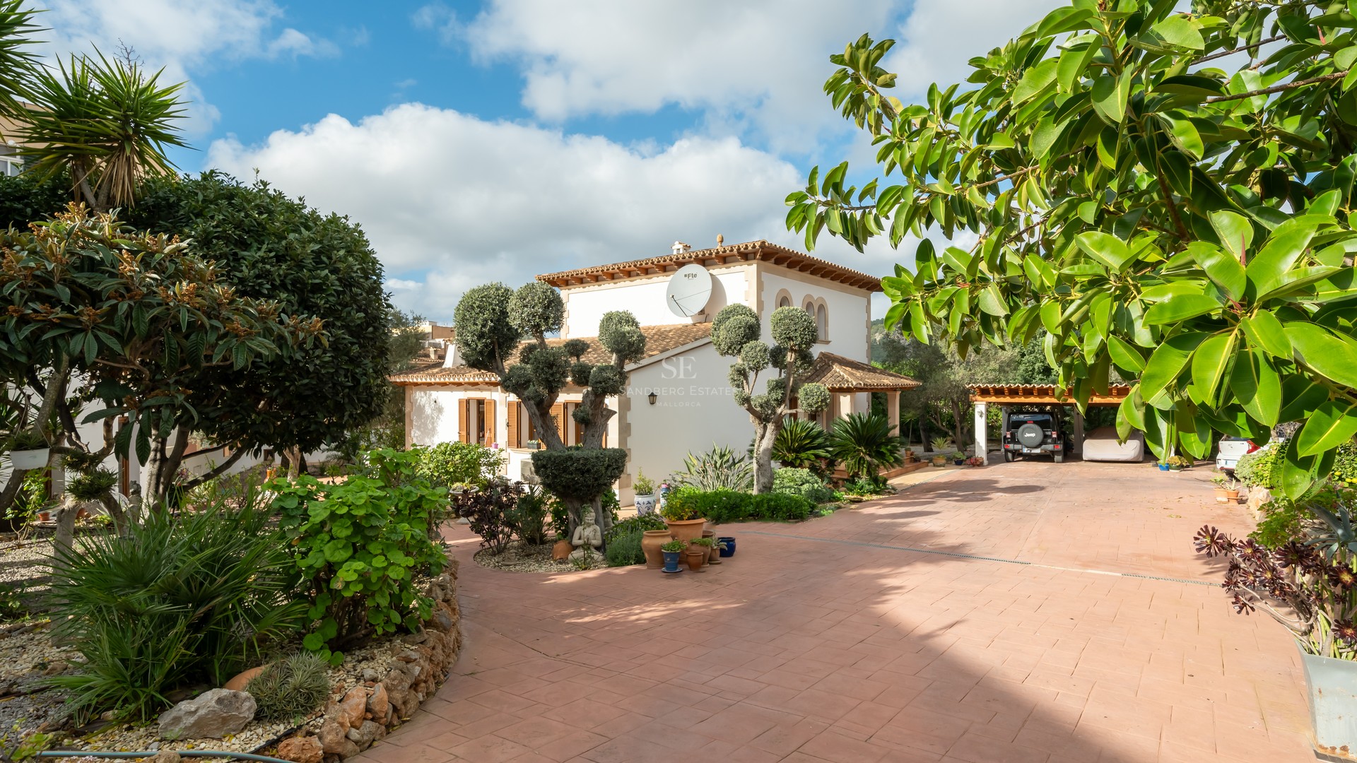 A sun-drenched Mediterranean villa with a terracotta driveway, manicured trees, and a covered carport.