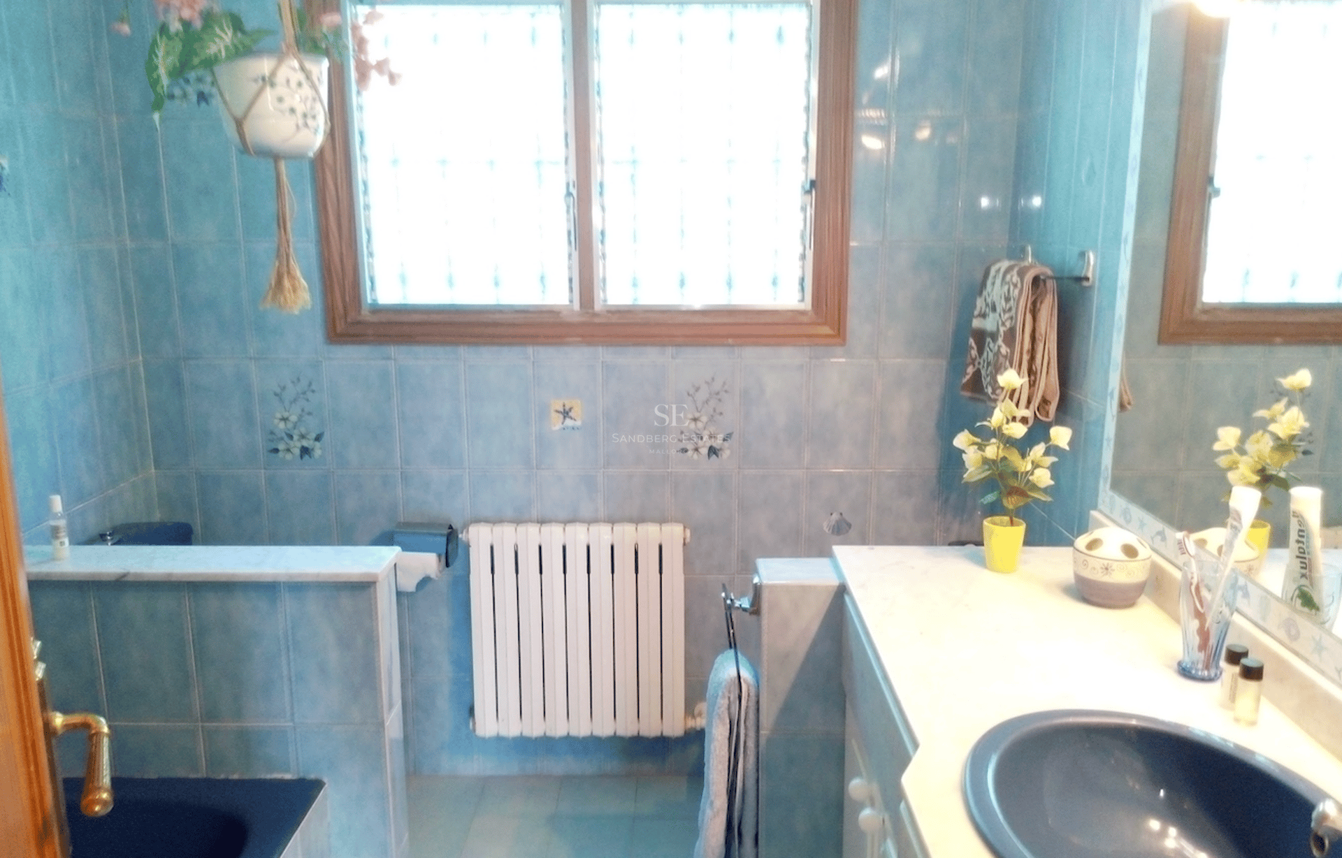 Baño con azulejos azules decorados, encimera de mármol blanco, ventana de madera y radiador blanco.