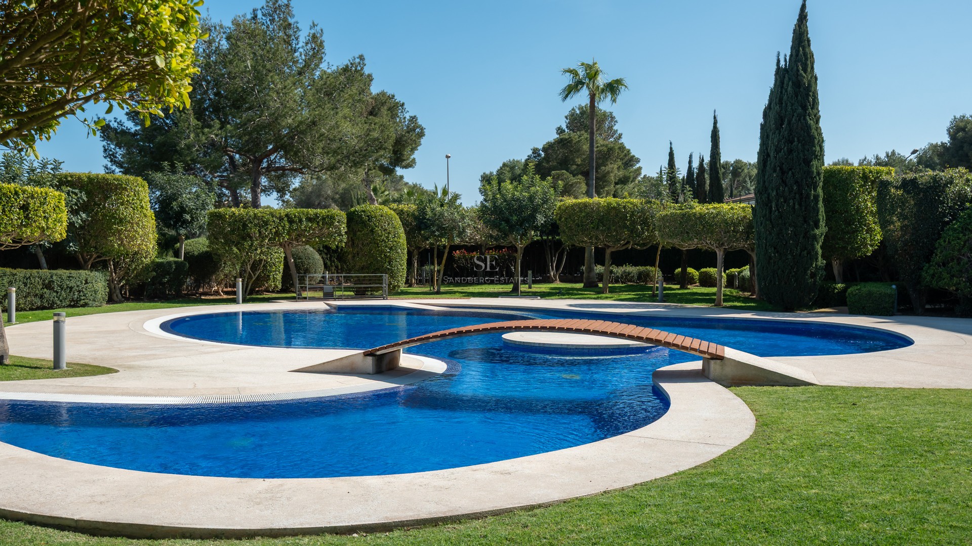 An expansive multi-lobed blue swimming pool with a wooden footbridge, surrounded by manicured lawns and mature trees.