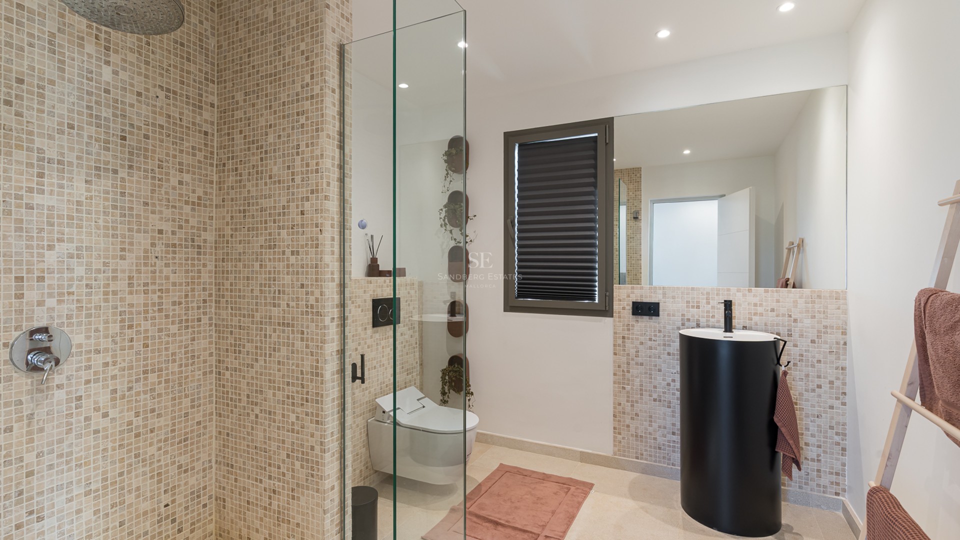 Modernt badrum med mosaikkakel, duschvägg i glas, mattsvart pelarhandfat och vägghängd toalett.