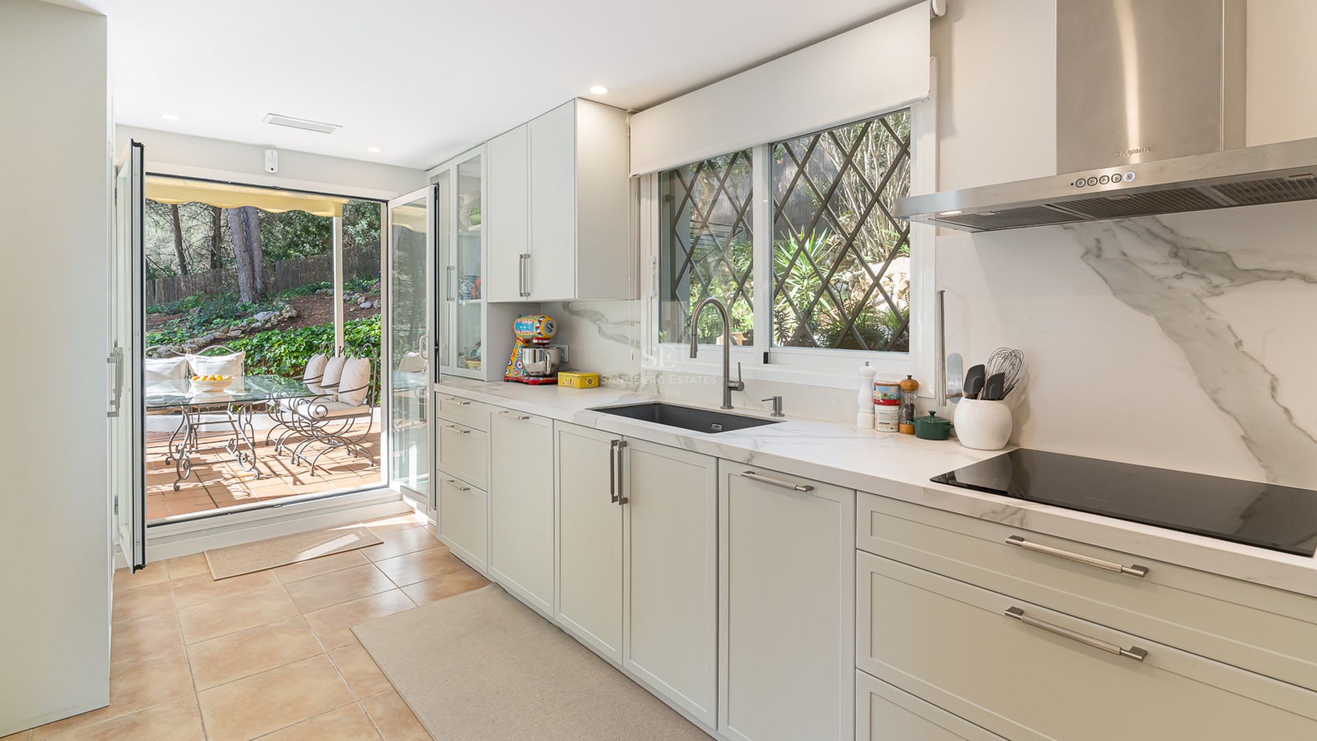 Bright kitchen with white cabinetry, marble countertops, and large glass doors opening to a lush garden terrace.