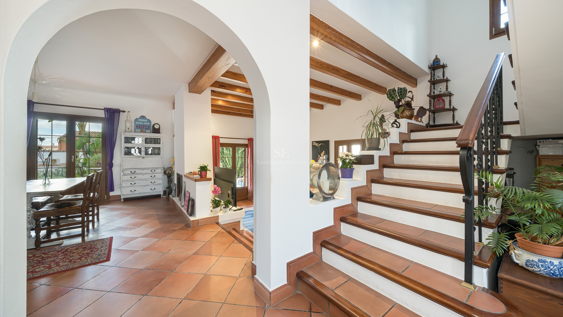Interior view showing terracotta floors, an arched entrance, wooden ceiling beams, and a staircase with iron railings.