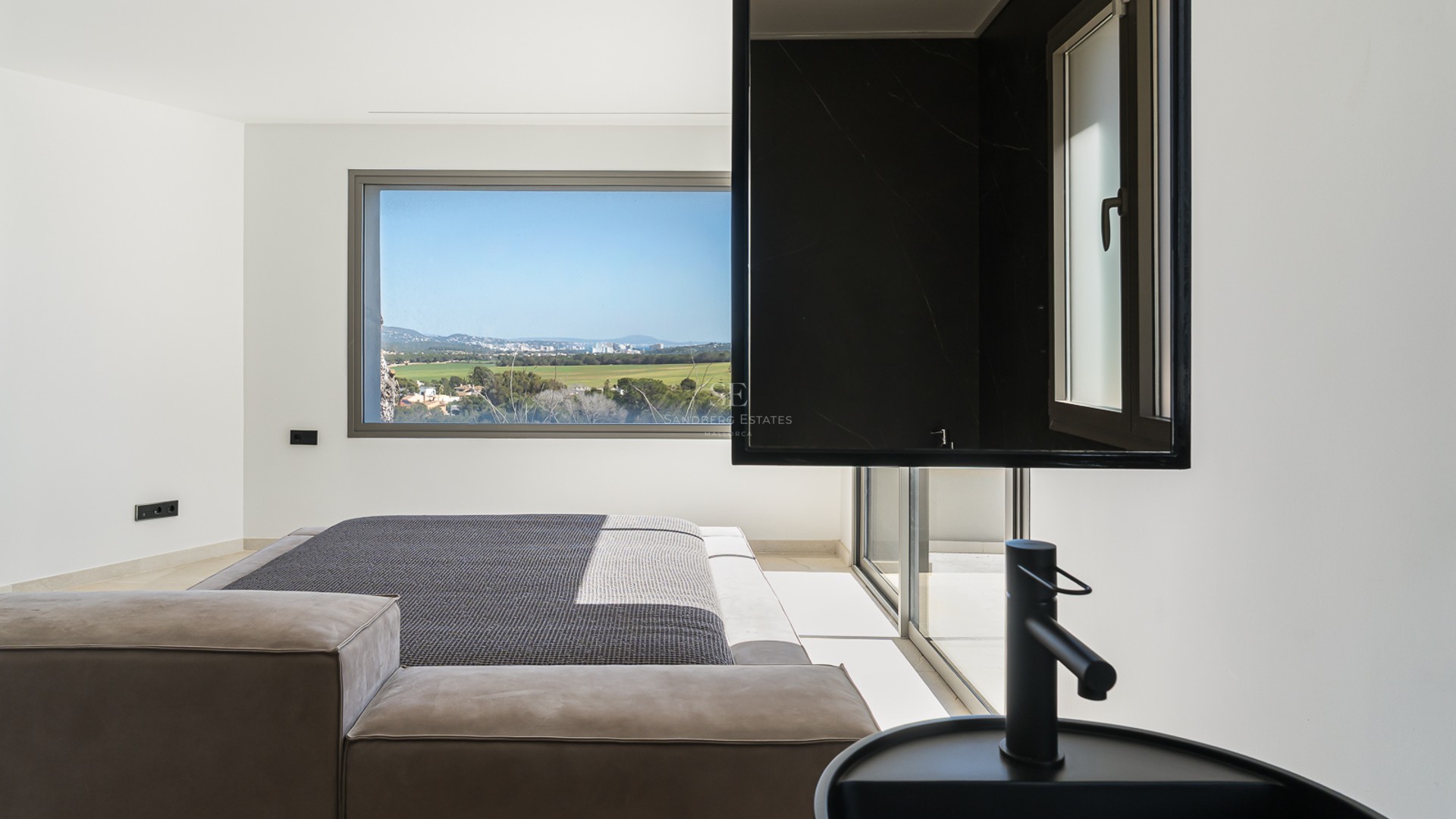 Modernt sovrum med integrerat svart tvättställ och ett stort fönster med utsikt över en grönskande dal.