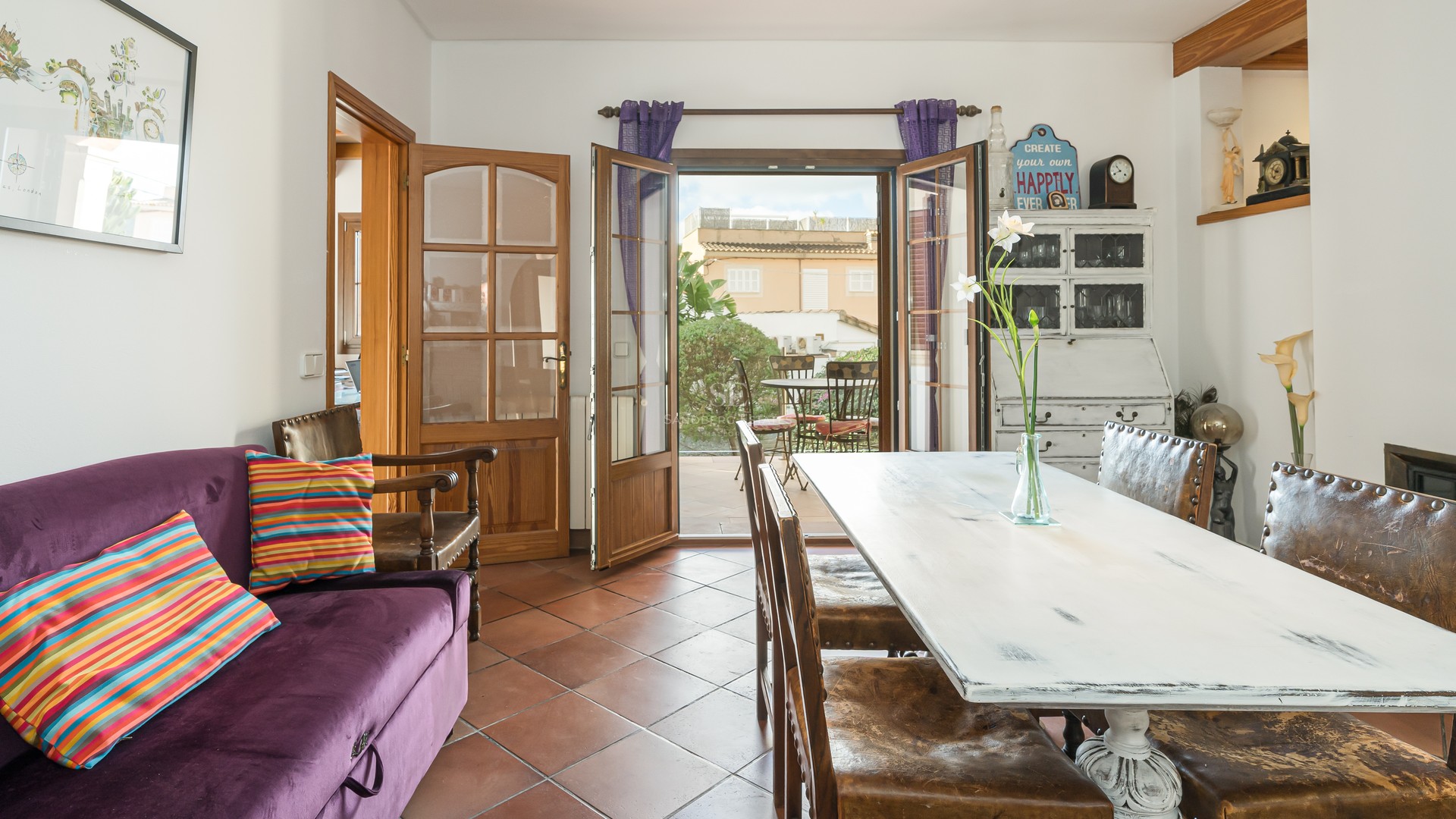 Interior dining room featuring terracotta tile floors, a rustic white wooden table, and open doors leading to a sunny patio.