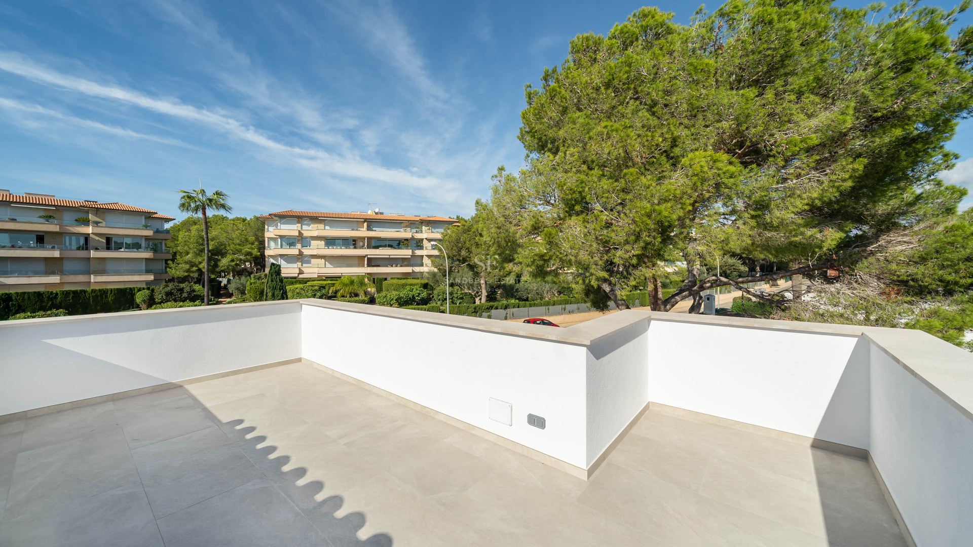 Grande terrasse moderne avec murs blancs et sol en pierre claire, donnant sur des pins luxuriants et des bâtiments.