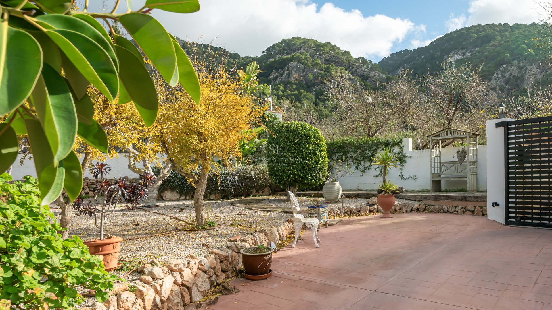 Terracotta terrace and landscaped garden with stone walls and mature trees against a rocky mountain backdrop.