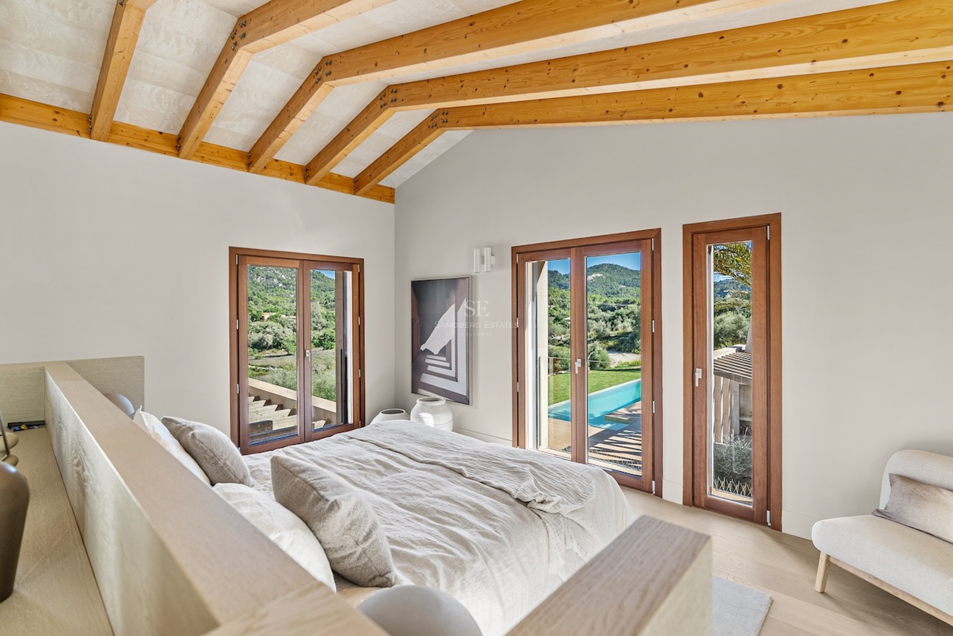 Chambre moderne avec plafond à poutres en bois, lit en lin beige et portes vitrées donnant sur une piscine et collines.