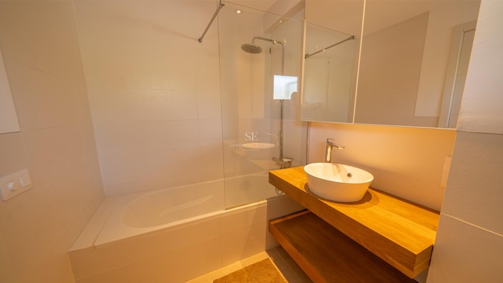 Baño minimalista con bañera blanca, mampara de cristal, ducha de efecto lluvia y mueble de madera con lavabo sobre encimera.