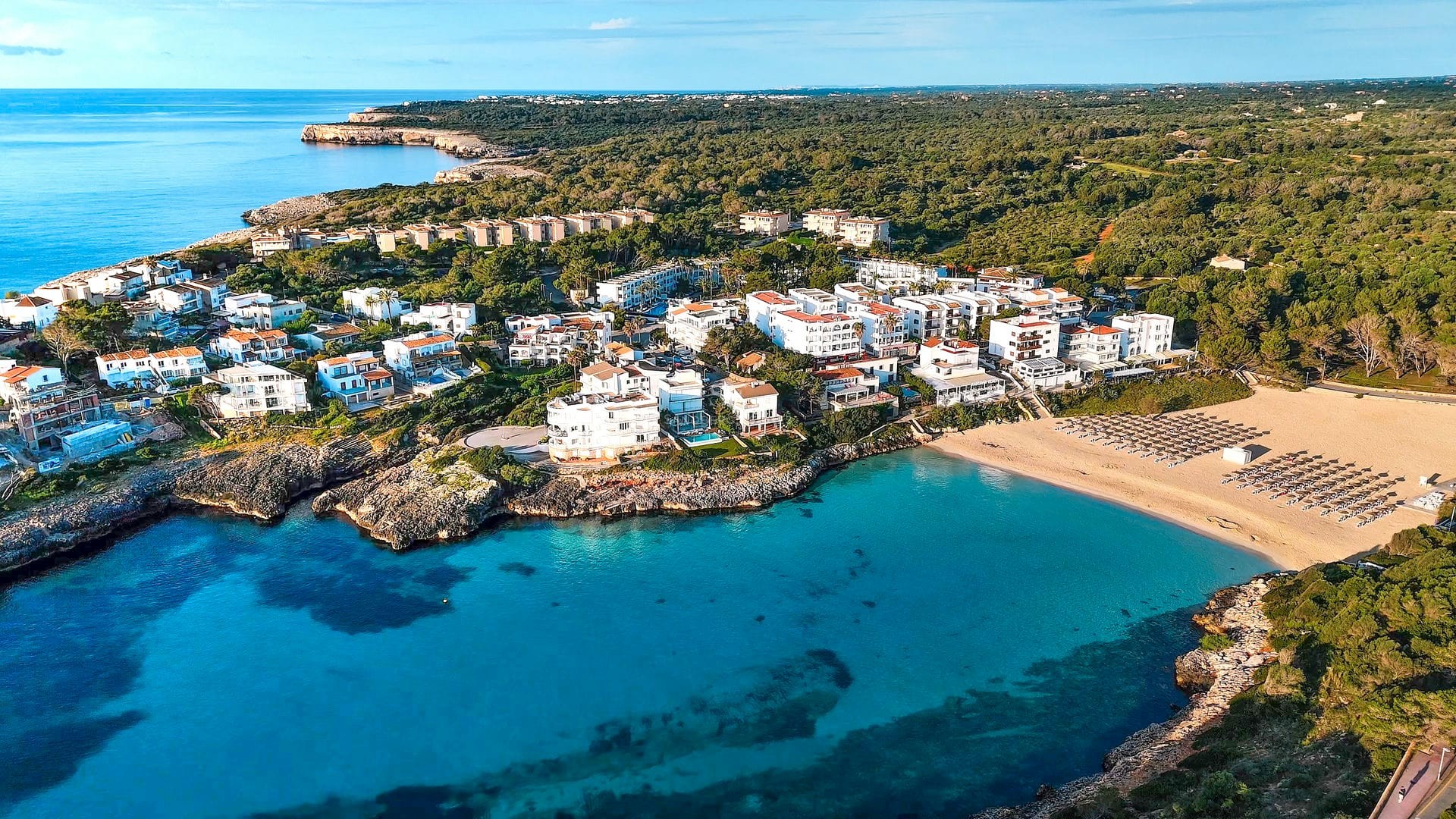 Vue aérienne d'un complexe de bâtiments blancs à côté d'une crique turquoise et d'une plage entourée d'une forêt verte.