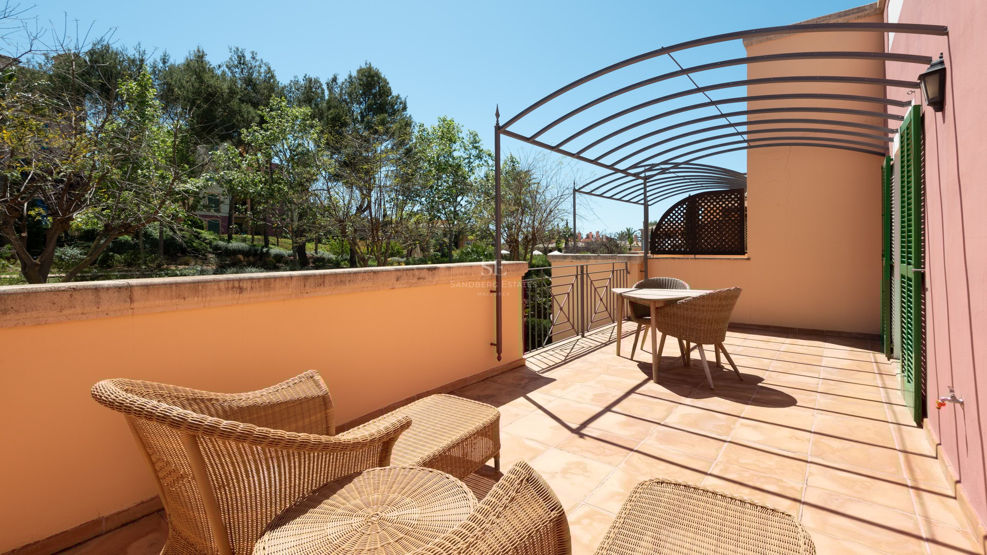 Spacious terrace featuring terracotta tiles, wicker furniture, and a metal pergola overlooking lush trees.
