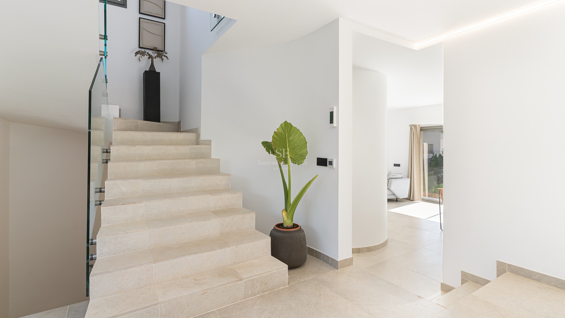 Modern inomhustrappa med trappsteg i ljus sten, glasräcken och en stor växt i en ljus entré.