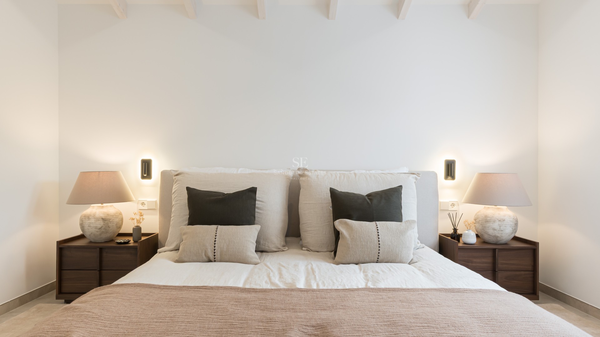 Lit avec tête de lit rembourrée, coussins décoratifs, tables de chevet en bois et lampes design à lumière chaude.