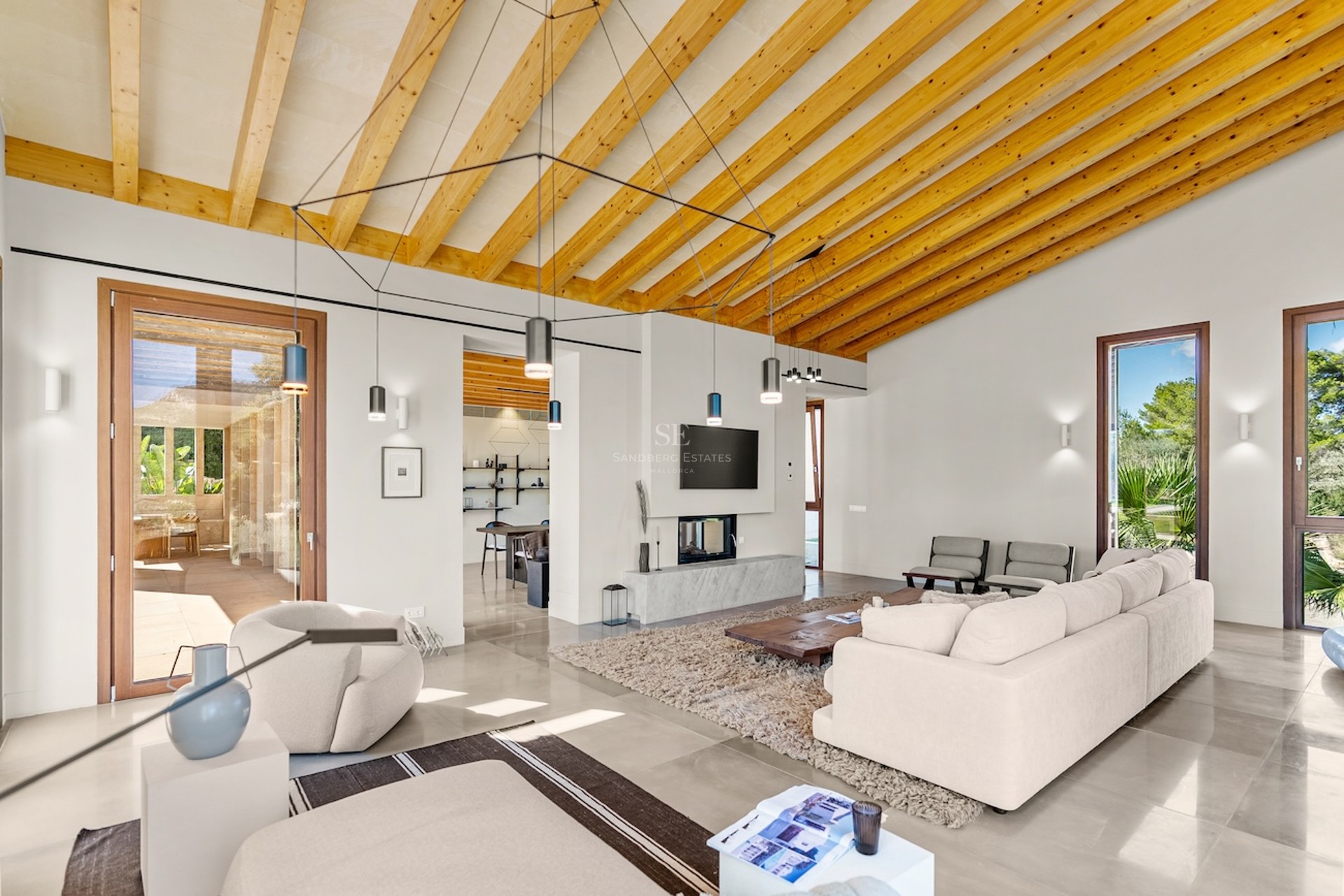 Salon lumineux avec plafond voûté en bois, canapés crème, cheminée minimaliste et grandes baies vitrées.