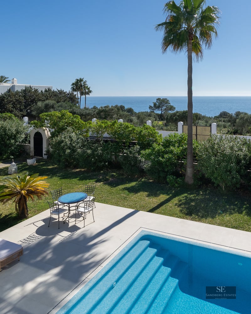 A sparkling blue swimming pool and patio set overlooking a lush garden and the Mediterranean Sea under a clear sky.