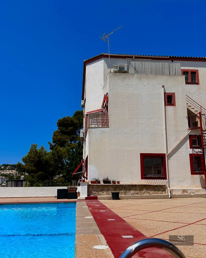 Large outdoor swimming pool in front of a white villa with red trim under a bright blue sky.