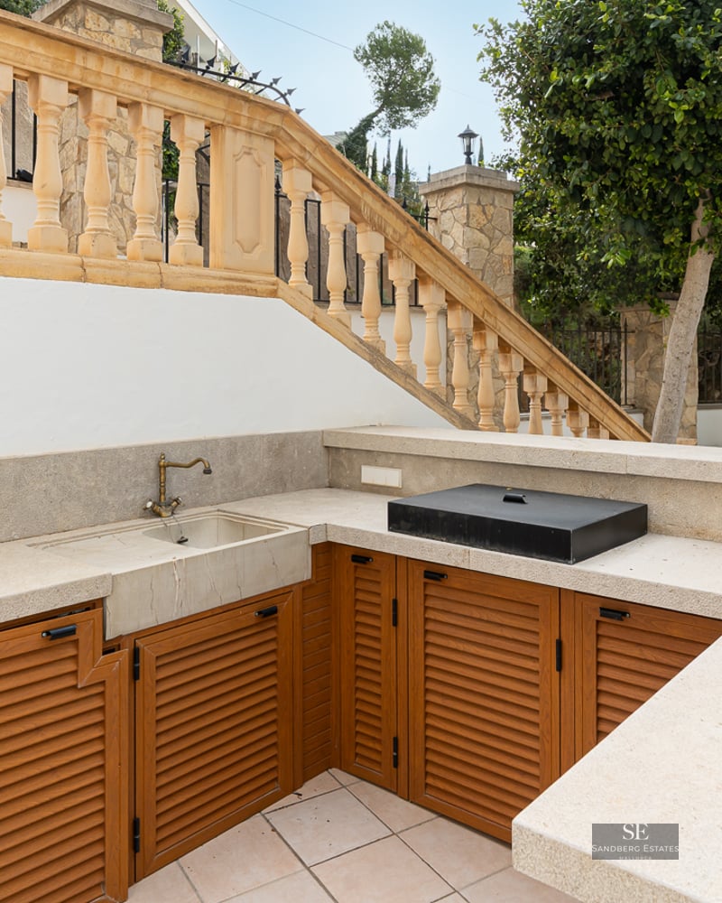 Outdoor kitchen with stone countertops, wooden cabinets, and a brass sink next to a classical stone balustrade.
