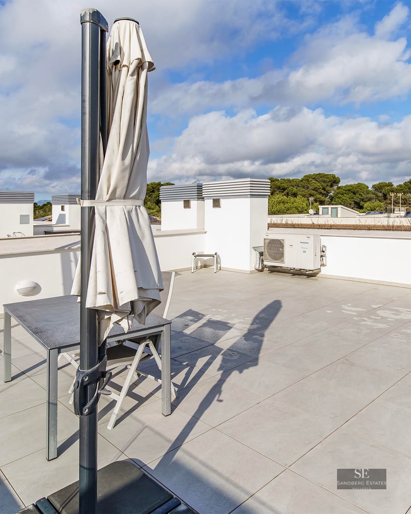 Spacious tiled rooftop terrace with grey table, chairs, and two closed white umbrellas under a blue cloudy sky.