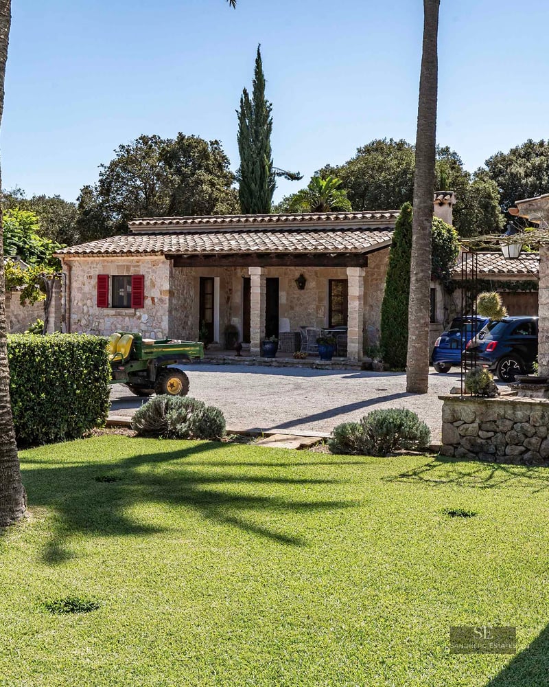 A rustic stone villa with red shutters and a terracotta roof, surrounded by a lush green lawn and palm trees.