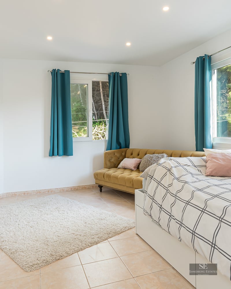 Chambre lumineuse avec lit, canapé en velours moutarde, rideaux bleu canard et vue sur la forêt par les fenêtres.