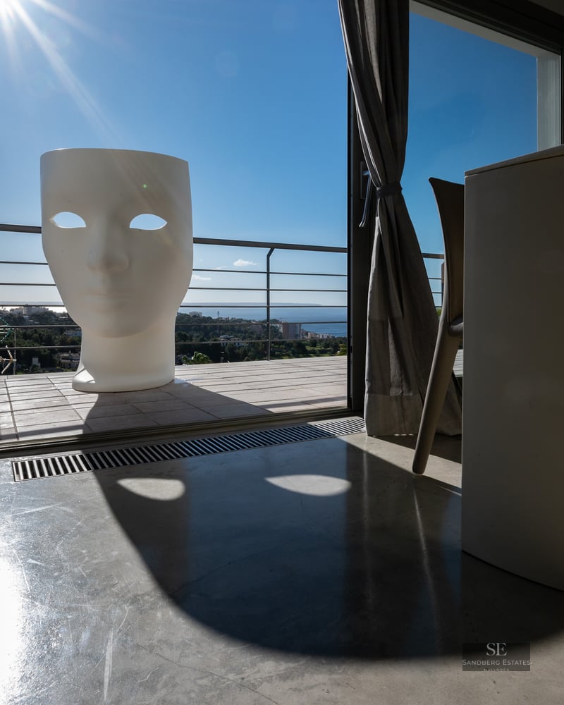 Interior view looking out to a sunny terrace featuring a giant white face-shaped chair and panoramic sea views.
