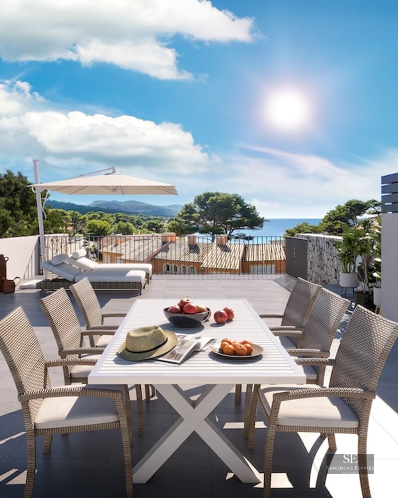 Sun-drenched rooftop terrace with white dining set, lounge chairs, and a panoramic view of the sea and coastal hills.