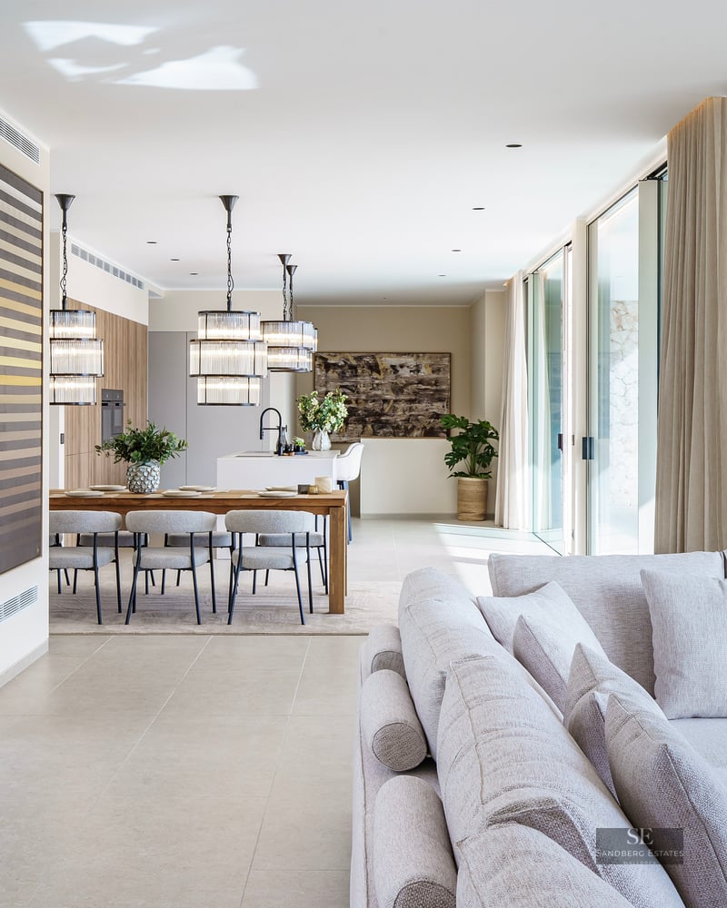 Spacious modern living room with light gray sofa, wooden dining table, and designer pendant lights by large glass doors.