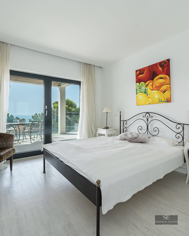 Bright white bedroom featuring a wrought-iron bed, animal print armchairs, and glass doors opening to a scenic sea-view terrace.