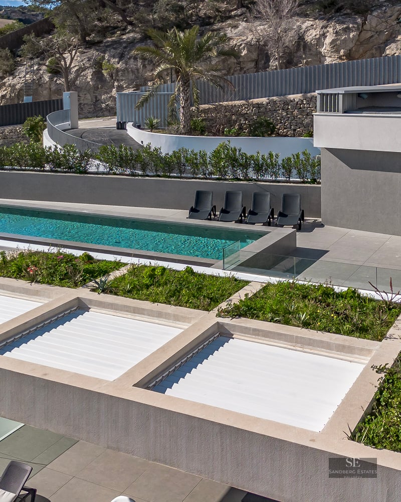 High-angle view of a modern grey villa featuring an infinity pool, sun loungers, and rooftop greenery against a rocky hill.