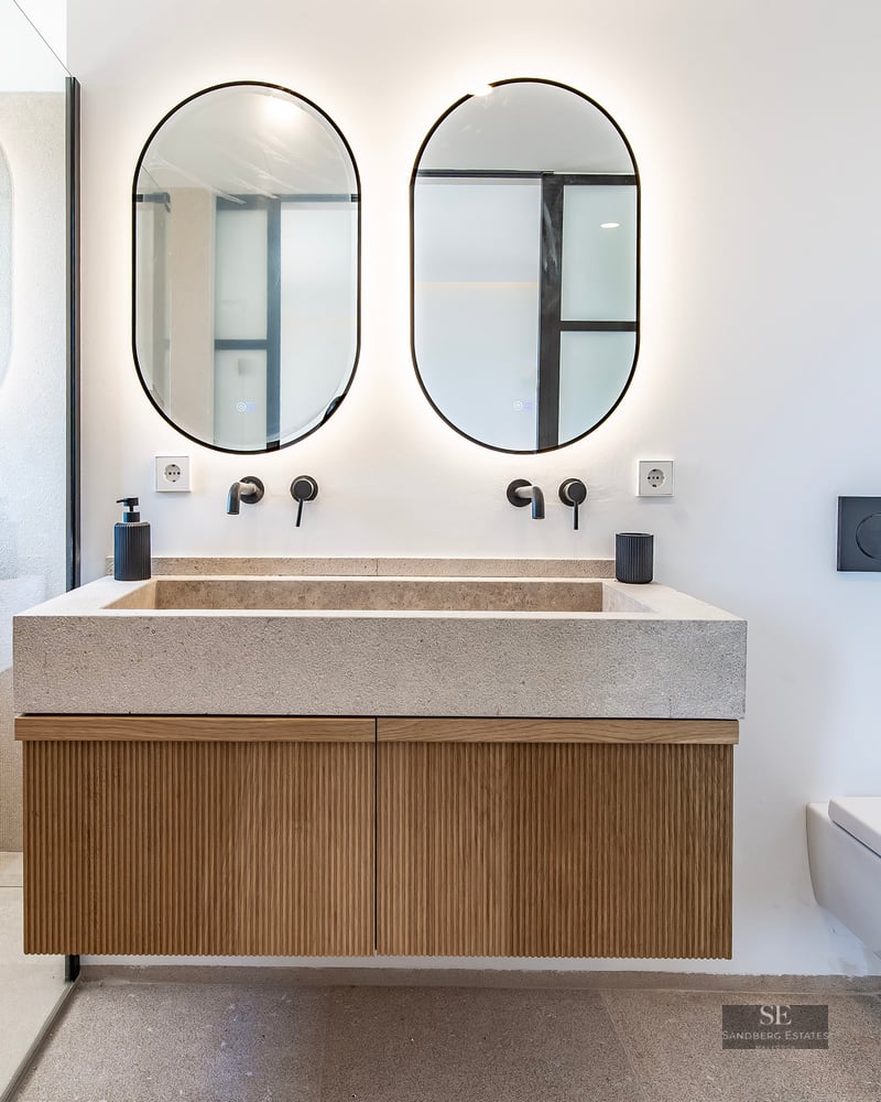 Modern bathroom featuring a stone double vanity, wooden cabinet, two oval backlit mirrors, and a walk-in glass shower.