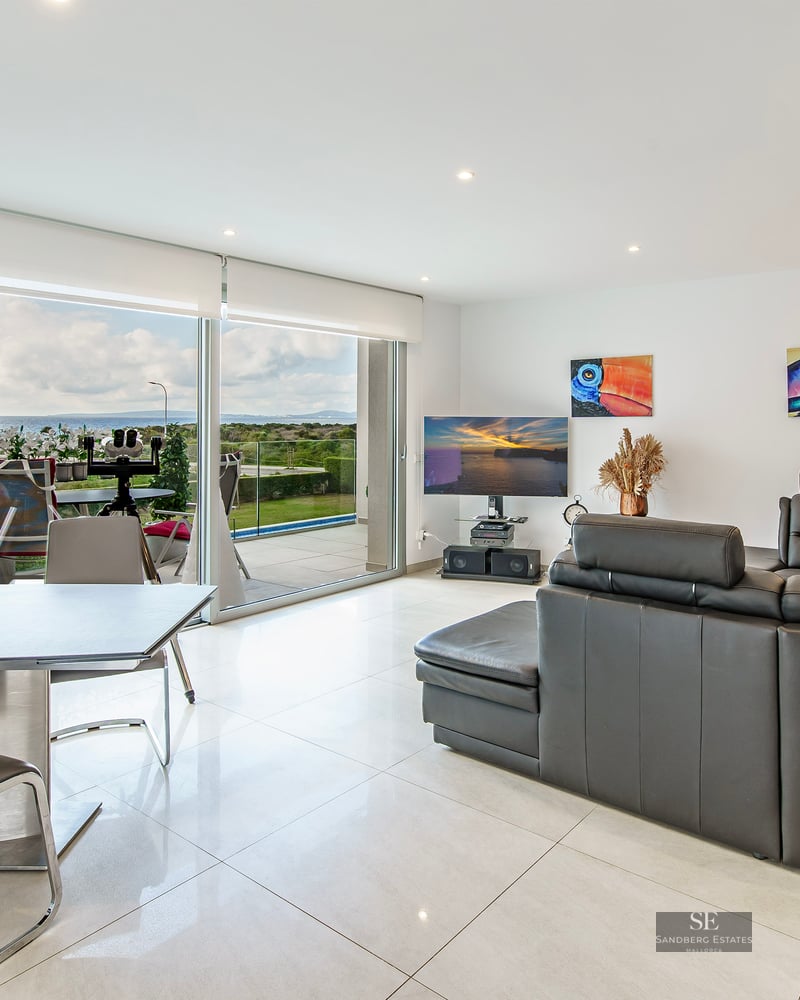 Modern living room with dark grey leather sofa, dining area, and large glass doors opening to a sea view.