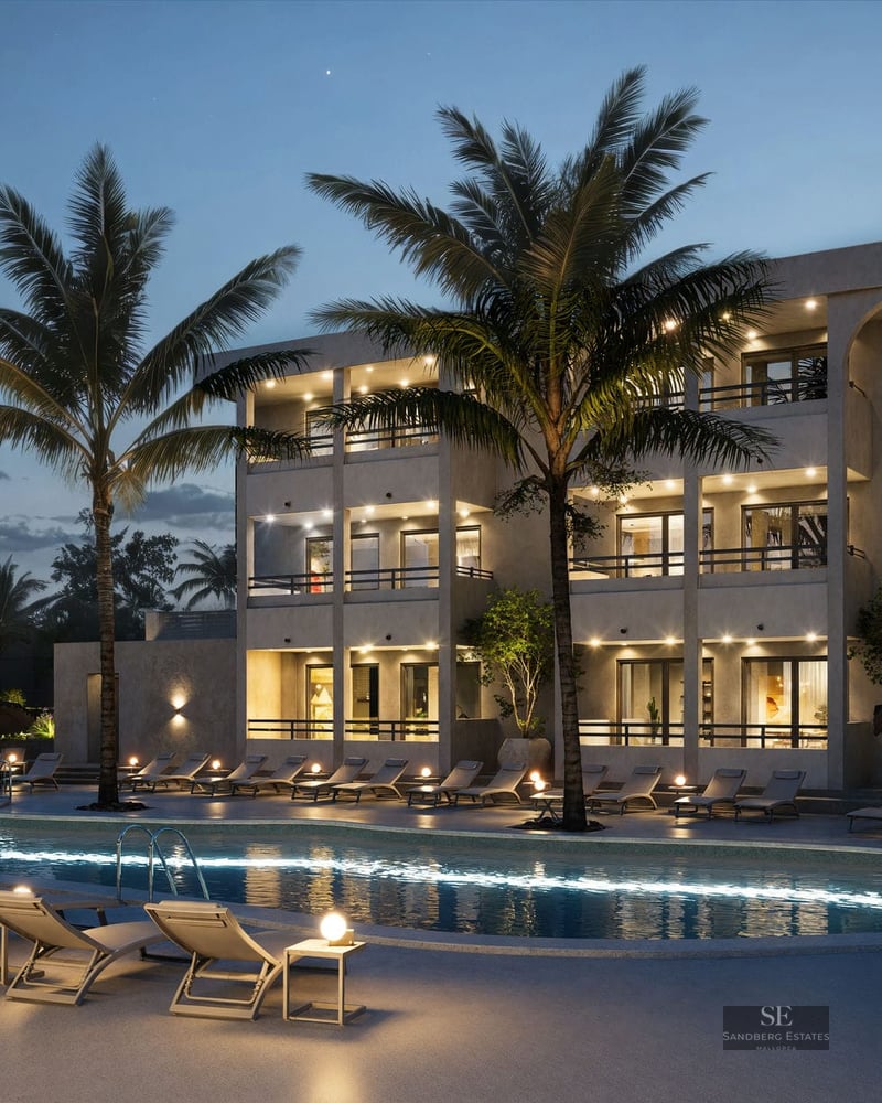 Grande piscine illuminée avec chaises longues et palmiers devant un immeuble d'appartements moderne au crépuscule.