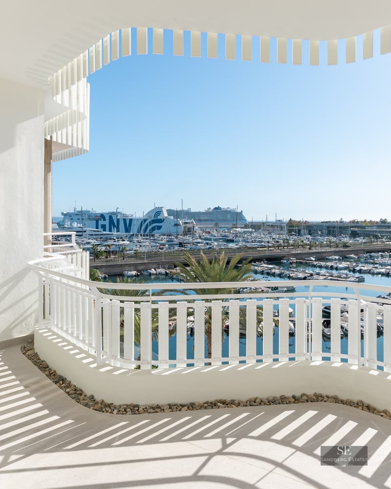 White curved balcony overlooking a sunny marina filled with boats and palm trees under a clear blue sky.