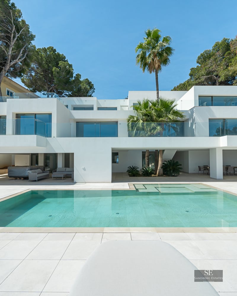 Modern white villa with a large turquoise pool, glass balconies, and palm trees under a clear blue sky.