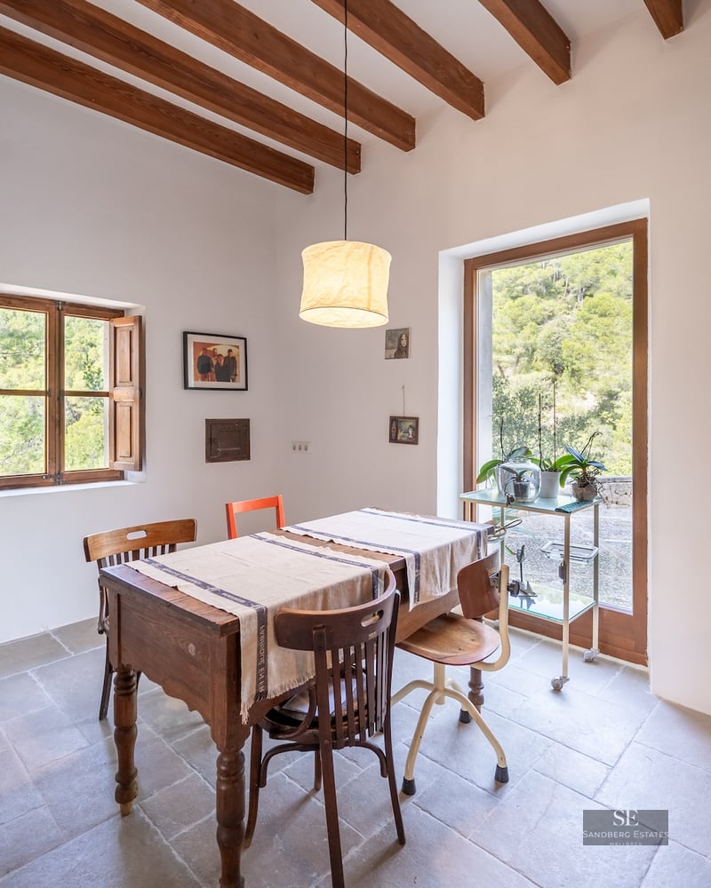 Bright dining room featuring a wooden table, mismatched chairs, exposed ceiling beams, and a glass door overlooking a forest.